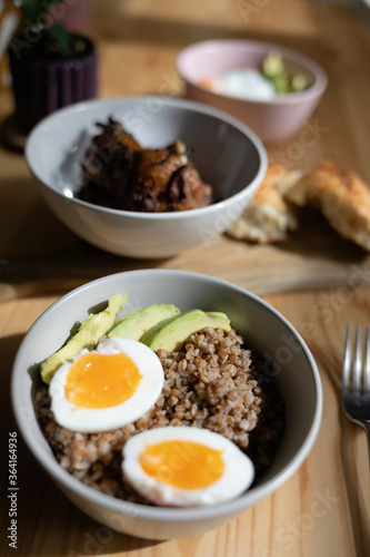 Buckwheat with boiled eggs and fresh green avocado in the bowl. Fried chicken and bread on a cutting board. Lunch over wooden with cutlery.