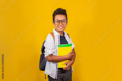 An African-American man stands on a yellow background with a backpack and a book smiling at the camera