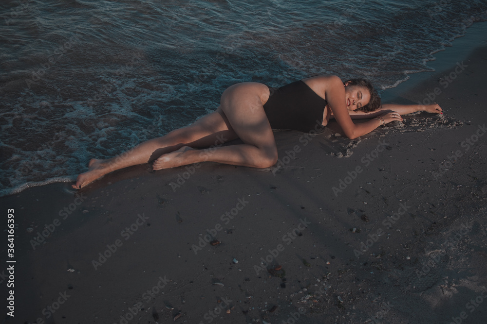 young brunette with long hair in a dark swimsuit lies on the sand on the shore with waves young brunette with long hair in a dark swimsuit lies on the sand on the shore with waves