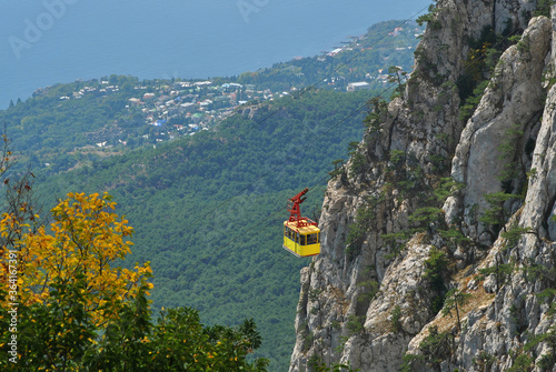The cable car goes up the mountain on a sunny day. Yellow cabin against the background of the mountains and the sea shore.