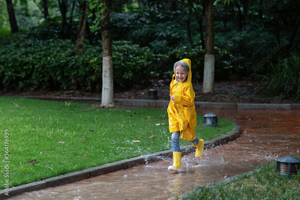 Cute little girl in yellow raincoat and rubber boots walking outdoor during rain. Bad weather, summer tropical storm, autumn fashion concept.
