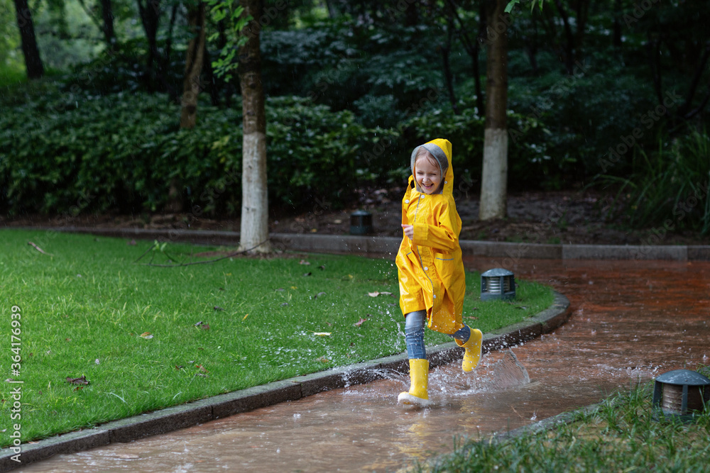 Cute little girl in yellow raincoat and rubber boots walking outdoor during rain. Bad weather, summer tropical storm, autumn fashion concept. Cute little girl in yellow raincoat and rubber boots walking outdoor during rain. Bad weather, summer tropical storm, autumn fashion concept.