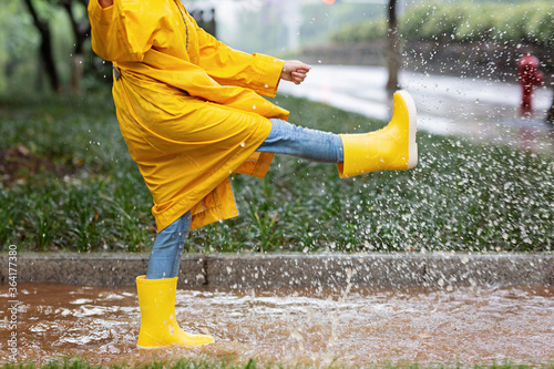 Cute little girl in yellow raincoat and rubber boots walking outdoor during rain. Bad weather, summer tropical storm, autumn fashion concept.