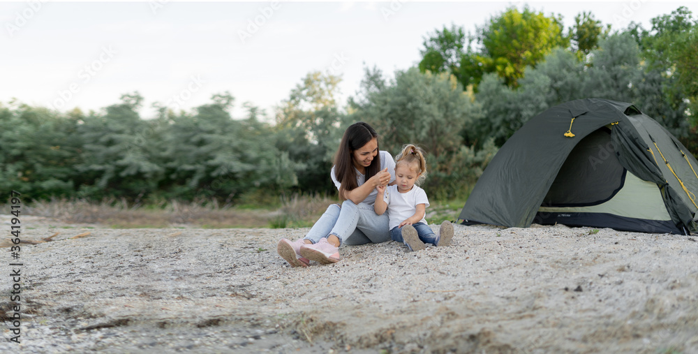 Smiling pretty young woman playing with her adorable daughter on the sandy beach. Hiking tent on the background.