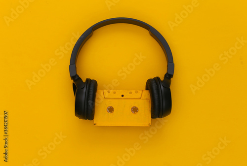 Black stereo headphones with a retro audio cassette on a yellow background. Top view
