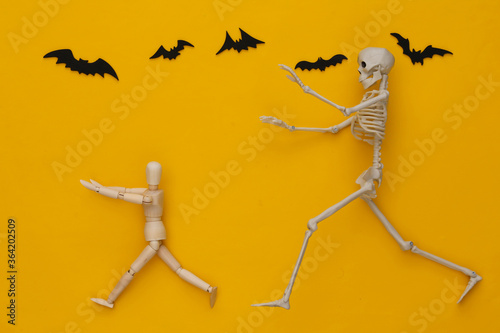 Halloween scary concept. Wooden puppet runs away from the skeleton on yellow background with flying bats