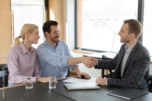 Excited young Caucasian couple handshake greeting with male real estate agent at meeting in office, happy husband shake hand close deal make agreement with broker, ownership, acquaintance concept