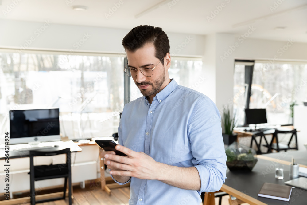 Young Caucasian male employee in glasses look on cellphone screen text or message in office, concentrated businessman use modern smartphone gadget, browse wireless internet at workplace