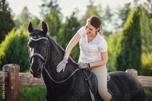 portrait of beautiful young woman and stunning friesian stallion in bridle and saddle standing near fence in summer