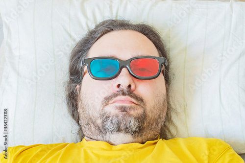 A middle-aged bearded man lies on a pillow in the early morning in colored 3D glasses. Concept - midlife crisis, childishness