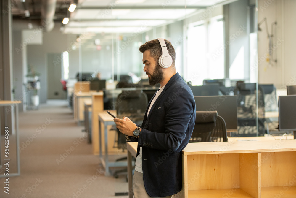 Contemporary young serious office worker with headphones scrolling in smartphone