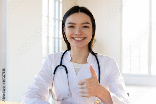 Head shot portrait smiling female doctor showing thumb up gesture, young woman wearing white coat uniform with stethoscope making video call, consulting patient online, good medical checkup result