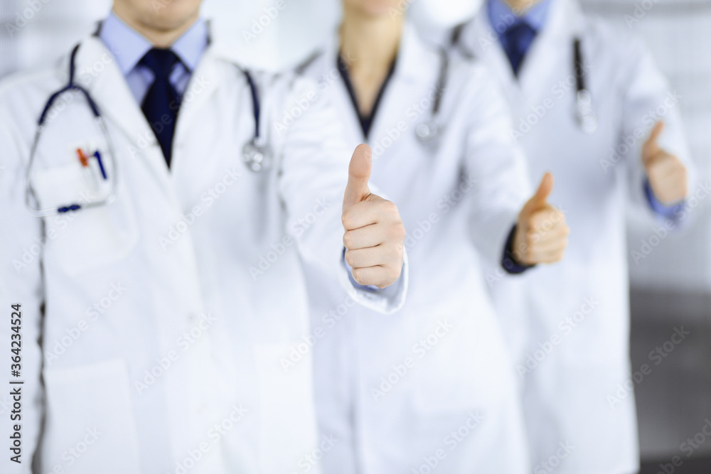 Group of unknown doctors stand as a team with thumbs up in a hospital office. Physicians ready to examine and help patients. Medical help, insurance in health care, best desease treatment and medicine Group of unknown doctors stand as a team with thumbs up in a hospital office. Physicians ready to examine and help patients. Medical help, insurance in health care, best desease treatment and medicine