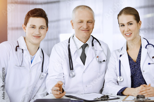 Doctors colleagues and elderly therapist discussing current disease therapy while sitting at working place in sunny clinic. Teamwork in medicine