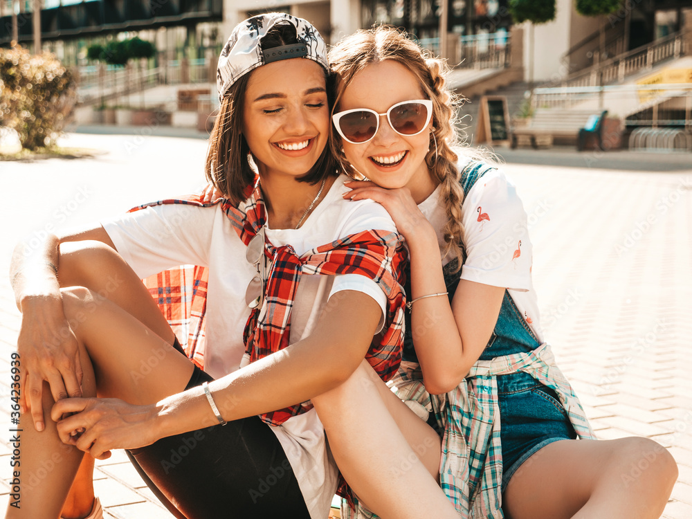 Two young smiling beautiful girls with colorful penny skateboards. Women in summer hipster clothes sitting in the street background. Positive models having fun and going crazy