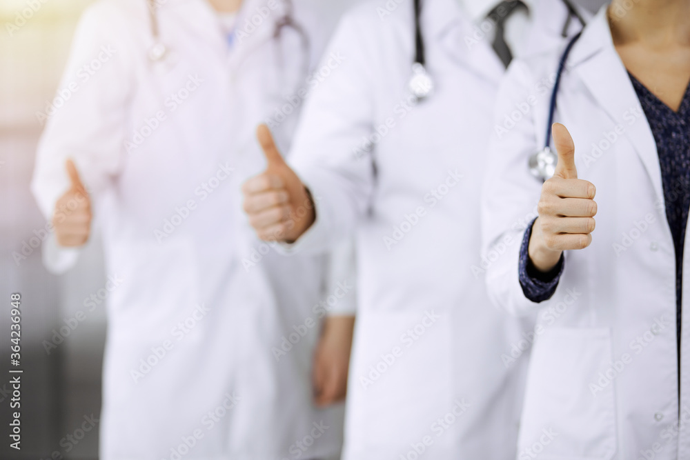 Doctors standing as a team with thumbs up in sunny clinic and ready to help patients. Medical help, insurance and medicine concept Doctors standing as a team with thumbs up in sunny clinic and ready to help patients. Medical help, insurance and medicine concept