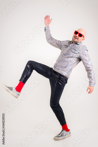 Strange caucasian man without hair in grey shirt, black leather trousers, black sunglasses, red socks and grey trainers isolated on white background