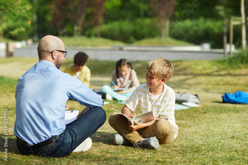 Full length portrait of smiling teenage boy sitting on green grass in sunlight and writing in notebook during outdoor class with teacher, copy space