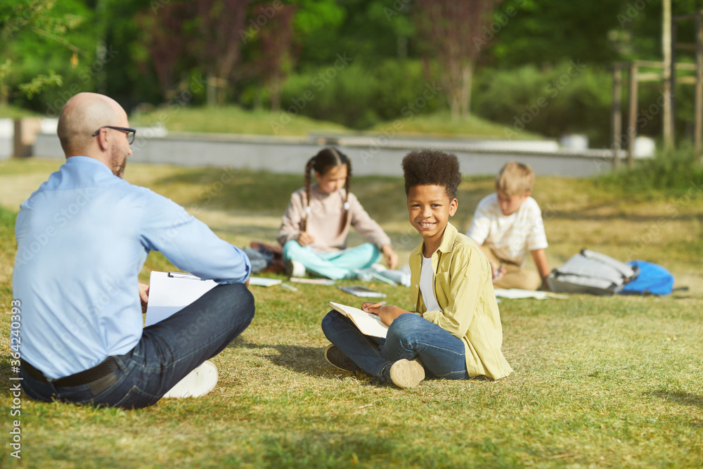 Full length portrait of smiling African-American boy sitting on green grass and looking at camera while enjoying outdoor class in sunlight, copy space Full length portrait of smiling African-American boy sitting on green grass and looking at camera while enjoying outdoor class in sunlight, copy space