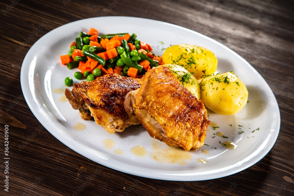 Roasted chicken thighs with boiled carrots, peas and potatoes on wooden background