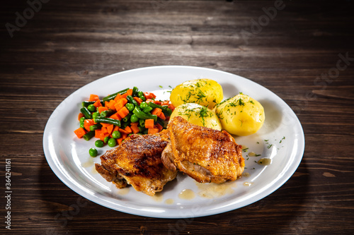 Roasted chicken thighs with boiled carrots, peas and potatoes on wooden background