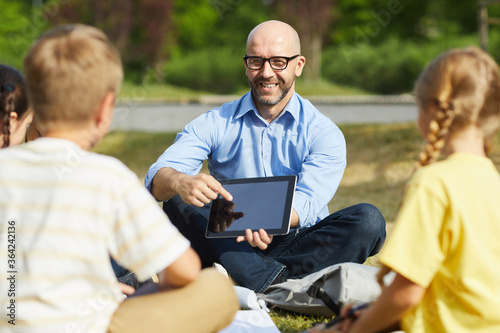 Portrait of bald male teacher pointing at tablet screen and smiling while talking to group of children during outdoor class in sunlight, copy space