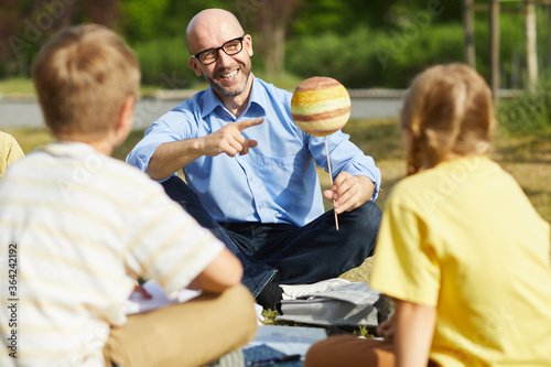 Portrait of bald male teacher pointing at planet model and smiling while talking to group of children during outdoor class in sunlight, copy space