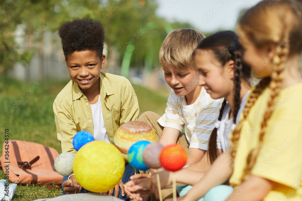 Fototapeta premium Multi-ethnic group of children sitting on green grass and holding model planets while enjoying outdoor astronomy lesson in sunlight, copy space