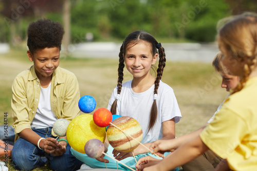Slika na platnu Multi-ethnic group of kids sitting on green grass and holding model planets whil