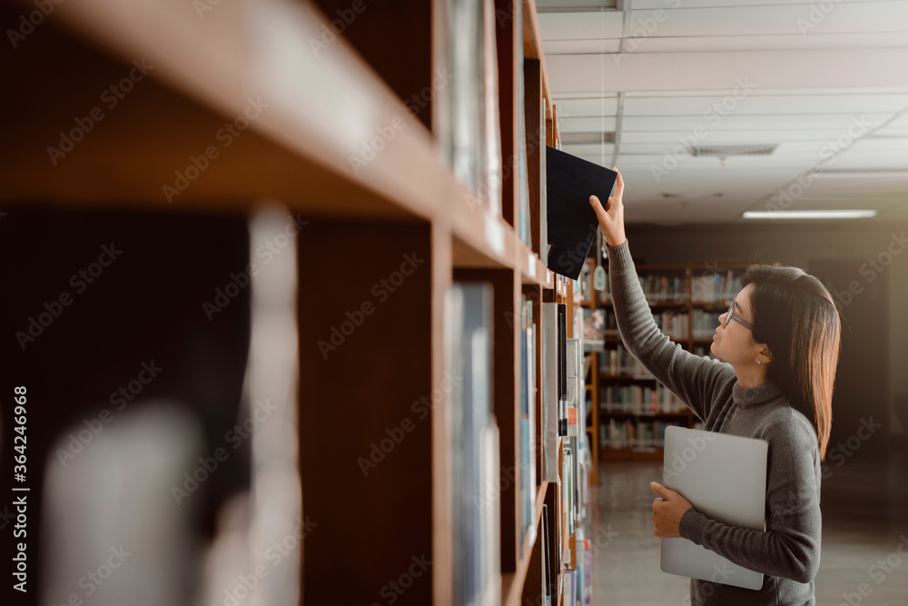 Close-up female hand picking book from bookshelf and holding a laptop in the library university surrounded by bookshelves with books. Close-up female hand picking book from bookshelf and holding a laptop in the library university surrounded by bookshelves with books.