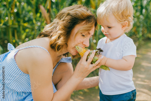 Picture of charming young caucasian woman with short hair in light blue dress relaxes with her little male baby with short fair hair in blue t-shirt and blue shorts in cornfield