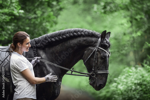 portrait of beautiful young woman standing near black friesian stallion in forest © vprotastchik