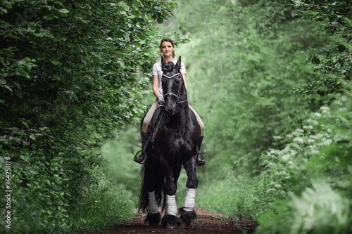 young woman rides black friesian stallion in forest