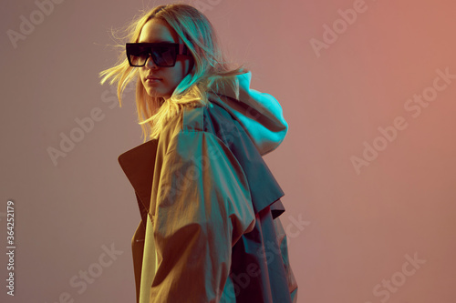 Stylish young girl, cool posing in a hoodie, sunglasses and cloak with developing hair, on a neon background.