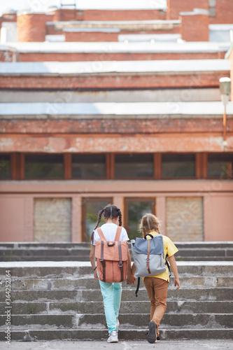 Vertical back view portrait of two sisters going to school with backpacks while walking up stairs to big building  copy space