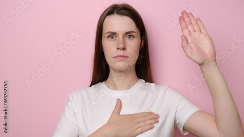 Portrait of sincere responsible young woman raising hand to promise, taking vow with serious dedicated expression, girl pledging allegiance, dressed in t-shirt,  isolated on pink studio background