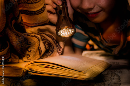 The child carefully reads the book by the light of a flashlight. The boy reads the story with interest. Cover your head with a blanket, a blanket.