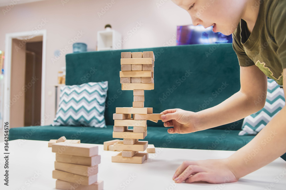 The boy builds a tower of blocks on the table. The game at the leaning tower. The child takes the cube out of the built tower so that it does not fall.