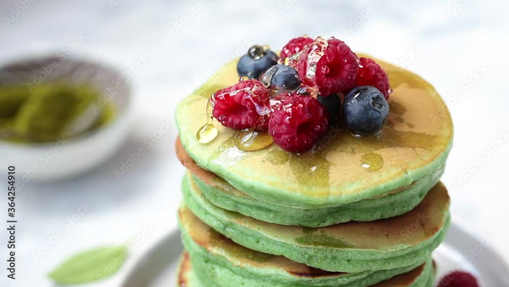 Pouring honey on green matcha pancakes with berries on a black plate.