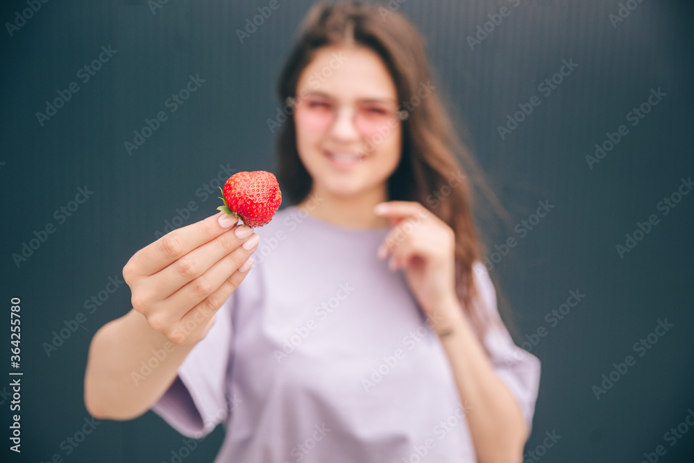 Young stylish trendy woman isolated over grey blue background. Blurred background of girl holding red strawberry in hand and smile. Casual woman posing on camera. Young stylish trendy woman isolated over grey blue background. Blurred background of girl holding red strawberry in hand and smile. Casual woman posing on camera.