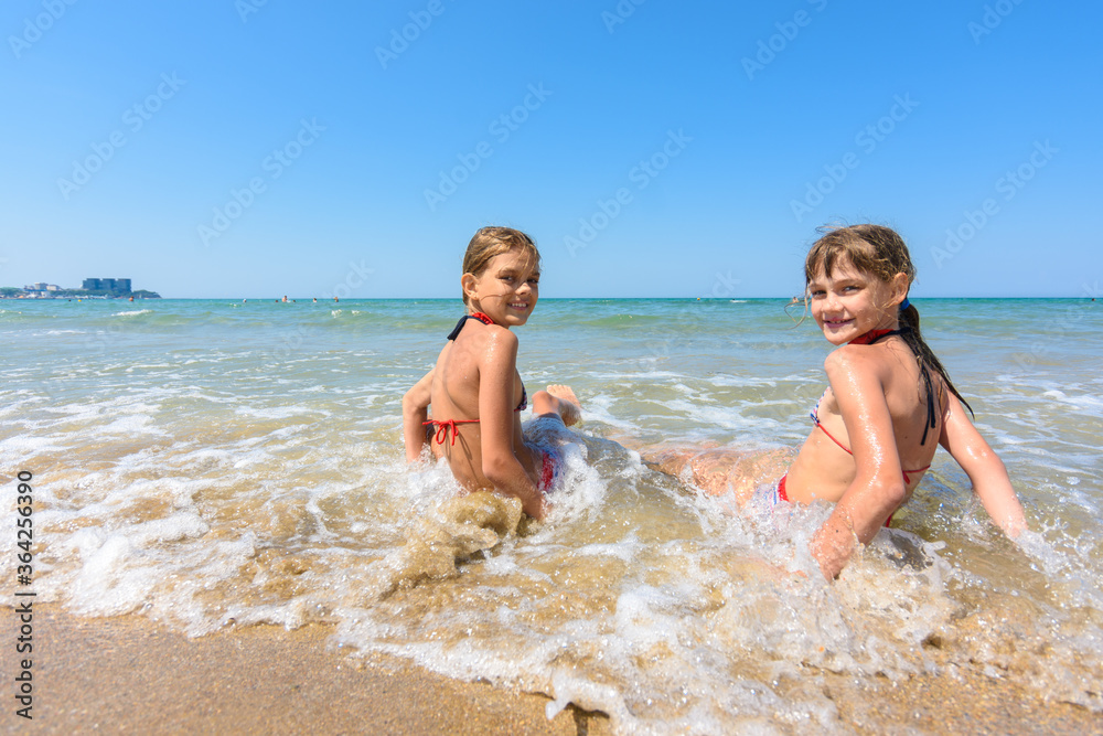 Two girls sit on the sea coast and turn around happily looking at the frame