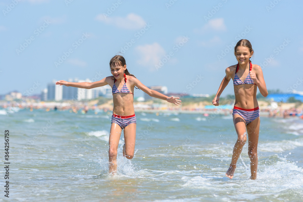 Two girls joyfully run along the seashore on a warm summer day