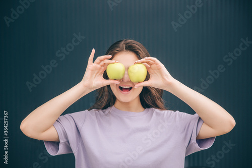 Young stylish trendy woman isolated over grey blue background. Funny positive girl covering eyes with tow ripe green white apples. Stand alone at dark wall. Healthy tasty fruit. © Vlad