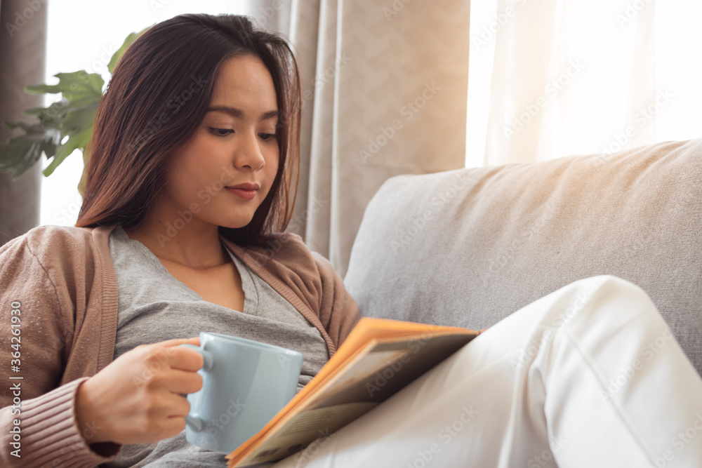 Young woman reading book and holding coffee cup on sofa. Young woman reading book and holding coffee cup on sofa.