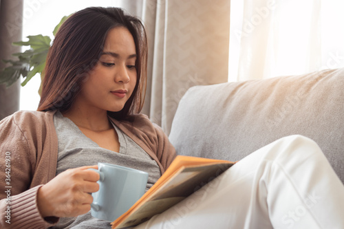 Young woman reading book and holding coffee cup on sofa.