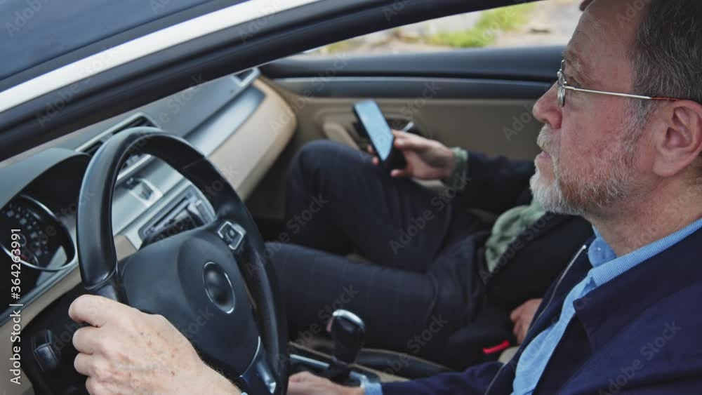 Old bearded man driving in the car for driver license test. Young instructor with smartphone giving directions. Business trip. Communication. City travel. Old bearded man driving in the car for driver license test. Young instructor with smartphone giving directions. Business trip. Communication. City travel.