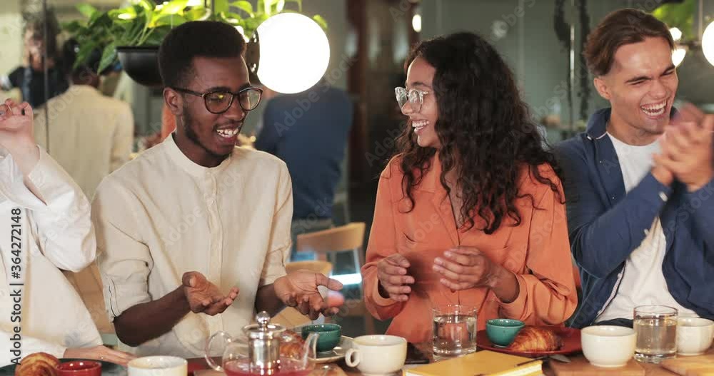Joyful african american young guy in glasses gesturing while telling funny story multiethnic friends in cafe. Happyl young colleagues listening and laughing. Concept of communication. Joyful african american young guy in glasses gesturing while telling funny story multiethnic friends in cafe. Happyl young colleagues listening and laughing. Concept of communication.