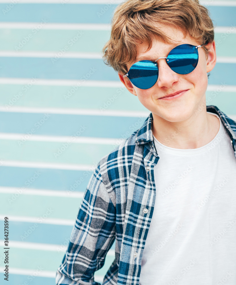 Blonde hair 12YO caucasian teenager boy Fashion portrait dressed white  t-shirt with checkered shirt in blue sunglasses with turquoise blue  background wall background. Teens fashion vertical concept. Stock Photo