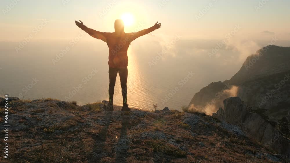 Filming from the back of an active man standing on the edge of the mountain and spreading his arms to the side from the amazing landscape of sunset and sea. Tourist male near a cliff looks at nature.