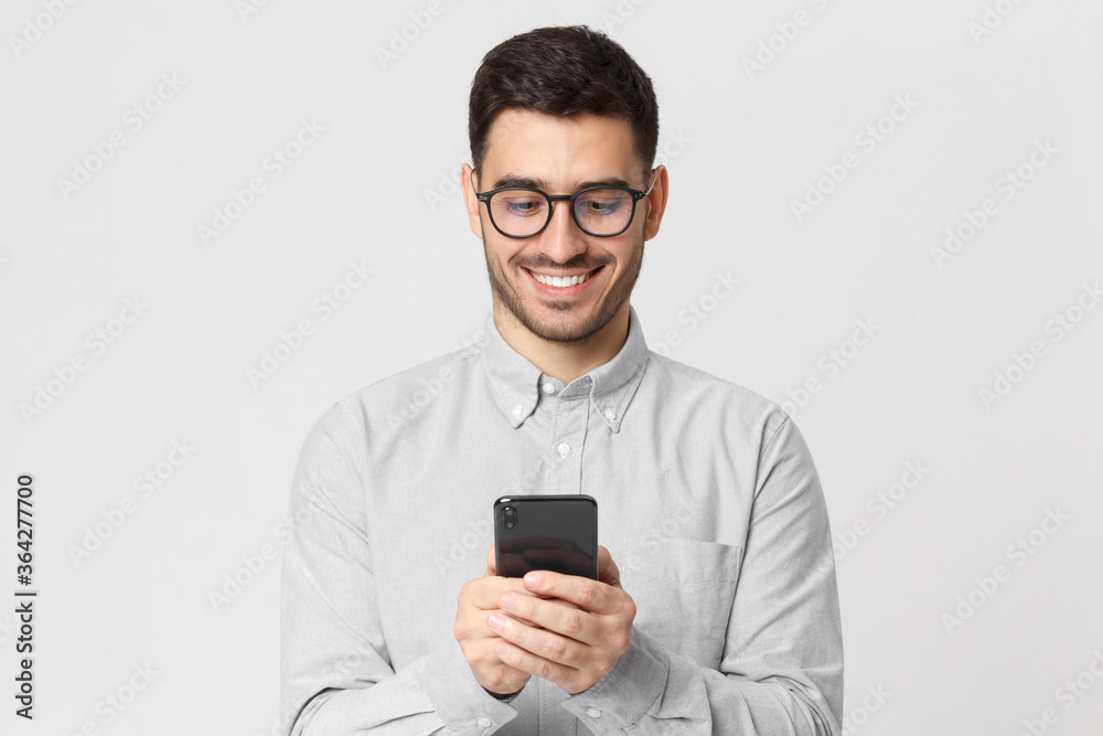 Young man wearing gray shirt and glasses, holding his smartphone and exchanging messages with friends with smile, isolated on studio background Young man wearing gray shirt and glasses, holding his smartphone and exchanging messages with friends with smile, isolated on studio background